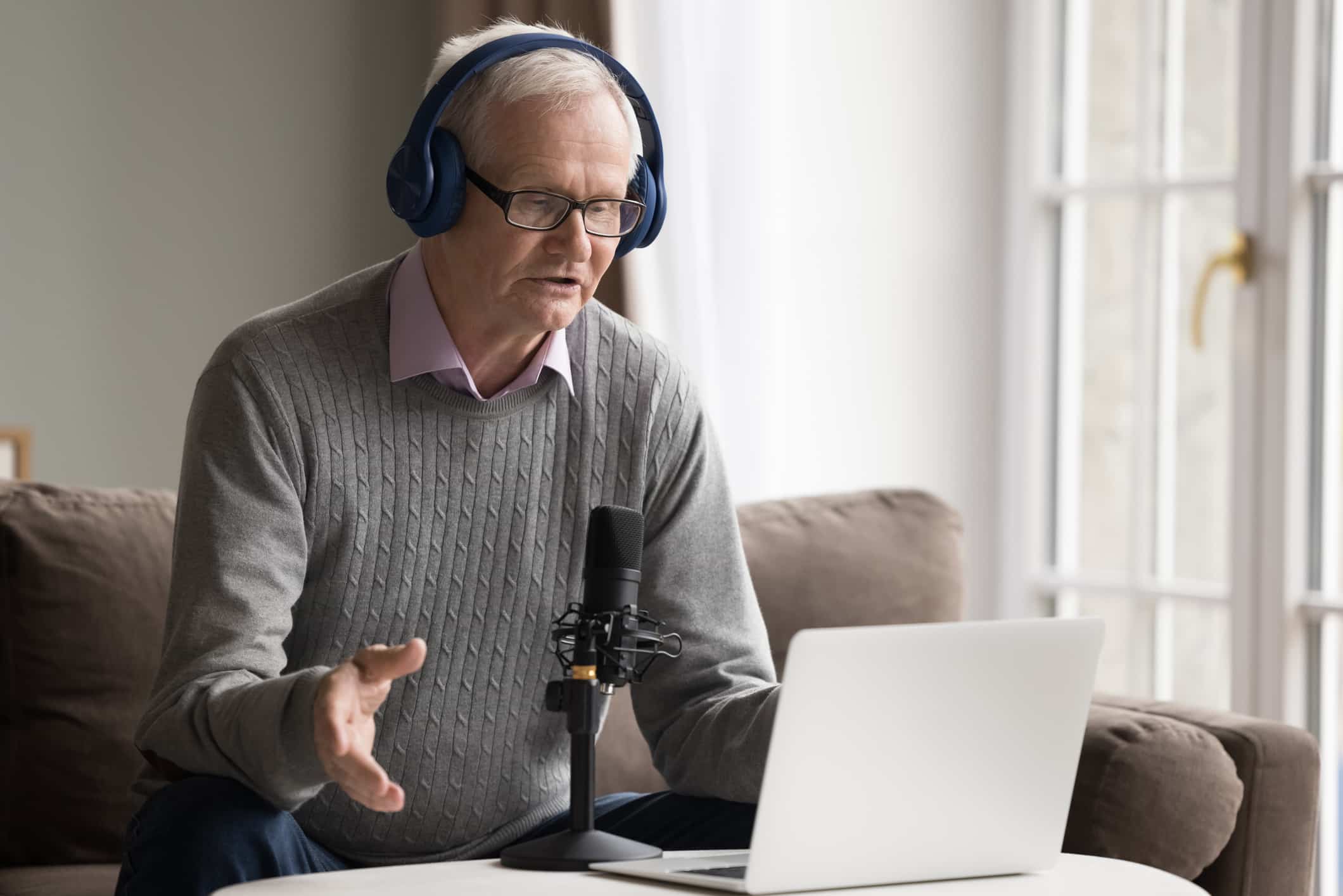 Older man in headphones makes speech into microphone. Serious older man in headphones lead webinar or stream for internet audience subscribers, makes speech into microphone look at laptop screen, provide information, participate in virtual event.